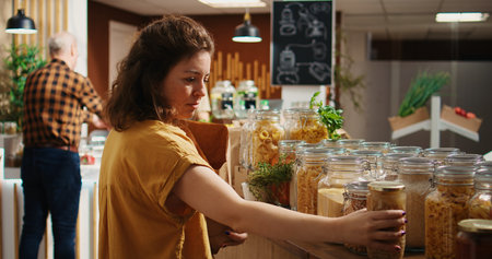 Happy woman in zero waste supermarket using paper bag to purchase pantry staples. Vegan customer in local neighborhood grocery shop doing shopping for cooking ingredientsの写真素材