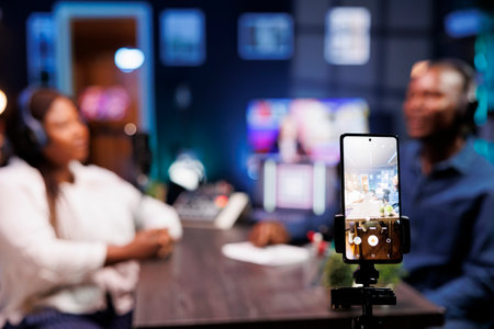 A vlogger is recording a podcast episode with a smartphone mounted on a tripod in a neon-lit studio. The show host is interviewing a guest and filming the discussion using a mobile device.の写真素材
