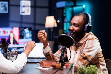 Smiling black man doing fist bump with african american presenter before starting live radio broadcast. Cheerful male influencer greeting guest during a recording session of online talk show.の写真素材