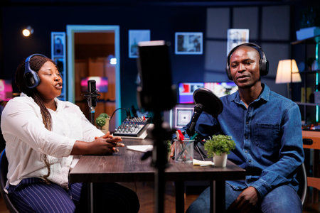 African American content creator couple talks tech in their home studio, entertaining a wide online audience with engaging discussions. Black vloggers using mobile device for recording radio program.の写真素材