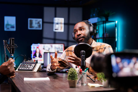 Talking with a guest, the show host uses a camera mounted on a tripod stabilizer to record a video for social media platforms. Male influencer using professional equipment to record a vlog at home.の写真素材