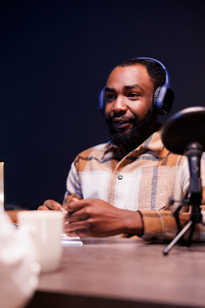 African american man using audio equipment to create an online radio show at home. Young black male content creator with wireless headphones and speaking into a microphone, is making a podcast.の写真素材