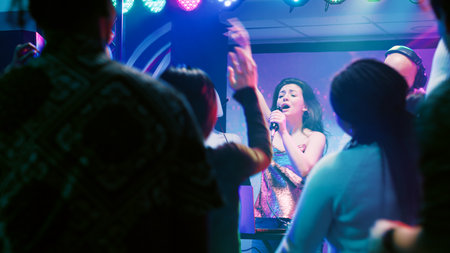 Confident woman doing karaoke on stage, singing songs with male DJ in front of people at nightclub....