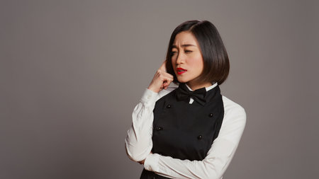 Asian waitress thinking about new menu ideas for restaurant, acting pensive in front of camera. Gourmet employee being creative with food plates, thoughtful server with apron and bow. Camera B.の写真素材