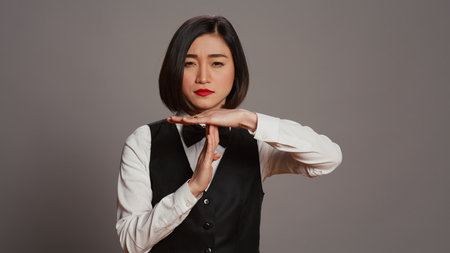Asian receptionist expressing timeout symbol on camera, showing negativity and asking for a work break. Tired bored employee showing pause sign in studio over grey background. Camera B.の写真素材