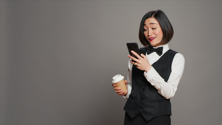 Concierge personnel drinking cup of coffee in studio, browsing smartphone social media apps. Asian receptionist enjoying caffeine refreshment while she uses mobile phone. Camera A.の写真素材