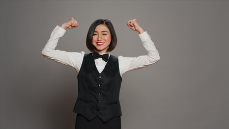 Asian hotel concierge flexing arms muscles in grey studio, showing off her strength and power in front of camera. Receptionist with bow and uniform standing with biceps flexed. Camera A.の写真素材