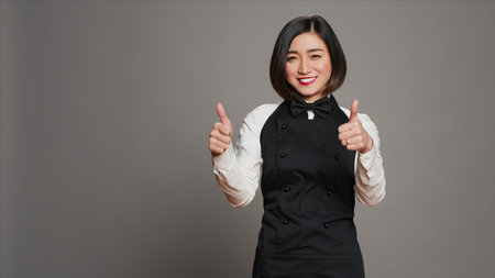 Smiling waitress giving thumbs up sign in front of camera, working in a five star restaurant to serve tables. Asian server with apron showing approval with okay symbol in studio, agreement. Camera A.の写真素材