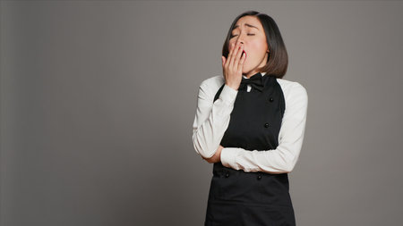 Exhausted waitress with apron yawning over grey background, feeling overworked and dealing with burnout after multiple restaurant events. Asian catering worker feeling tired in studio. Camera A.の写真素材