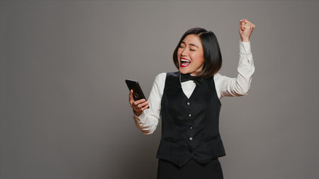 Front desk staff in uniform looks at online information on mobile phone, checking all website reservations over grey background. Asian receptionist using smartphone for bookings. Camera A.の写真素材