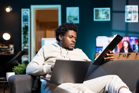 Remote employee concentrating on finishing up important job tasks, reading files info in apartment personal office. Focused teleworker crosschecking documents between laptop and paperworkの写真素材