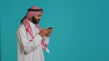 Happy person having fun with game on his smartphone device, wearing traditional arabic attire. In the studio, muslim model enjoying e sport tournament with mobile phone videogames.の写真素材
