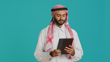 Middle eastern guy browsing tablet on camera, navigating webpages online on digital device. Male model in arabic clothing using gadget to check internet sites, texting on social media.の写真素材