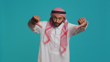 Arab adult with national gown and kufiyah shows thumbs down and feels dissatisfied with news. Young man expresses an apparent denial and negative gesture on camera, arabic person.の写真素材