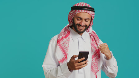 Arab guy holding his mobile device, surfing across social media platforms and chatting online. Muslim with customary apparel uses smartphone to access global web, feeling excited on camera.の写真素材