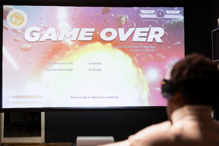 Gamer wearing headphones upset at seeing game over message on widescreen TV while playing arcade space shooter videogame. Man relaxing at home on gaming system, disappointed after losingの写真素材