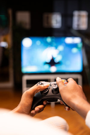 Focus shot on man holding controller in apartment, playing videogames on smart TV display in blurry...
