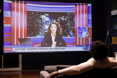 Bored man watching anchorwoman present breaking news while idling in living room. Cord cutter lounged on couch looking at online journalism channel streamed on widescreen smart TVの写真素材