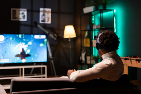 Man plays science fiction singleplayer game on gaming console while talking with friends through headphones. Player enjoying videogame with high quality graphics, using gamepad to navigate spacecraftの写真素材