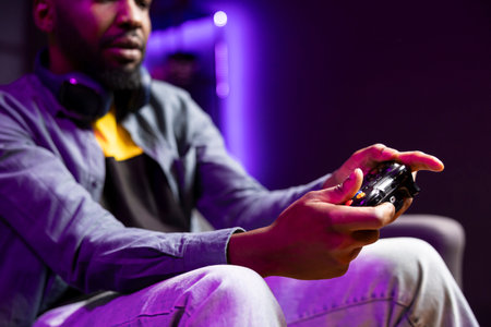 Gamer on couch using controller and headphones to play videogame at home, close up. African american man in home theatre using high tech gaming console gamepad to defeat opponents in gameの写真素材