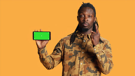 Cheerful adult holds smartphone with greenscreen in studio, presenting isolated copyspace display on mobile phone layout. Young person showing blank chromakey mockup on device.の写真素材