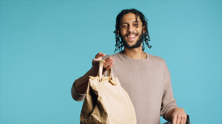 Portrait of pizza delivery guy taking takeaway paper lunchbag out of thermal bag. Middle Eastern courier getting meal out of food backpack, isolated over blue studio background, camera Aの写真素材
