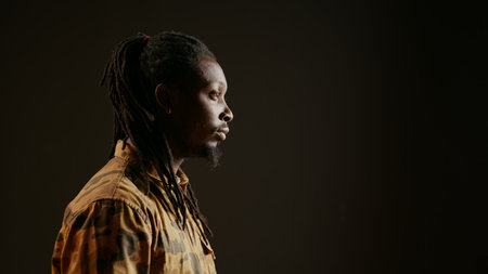 Smiling person posing in studio with black background, feeling confident wearing dreads and camo clothing. Cool guy with trendy hair acting stylish on camera, attractive man. Handheld shot.の写真素材
