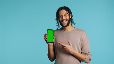 Excited man pointing index finger towards isolated screen phone, doing recommendation. Joyous person presenting mockup smartphone, doing positive gesturing, blue studio backgroundの写真素材