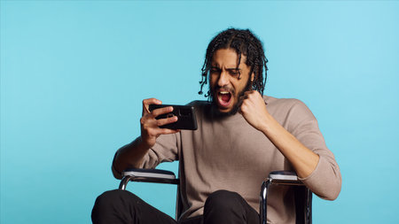 Joyful person in wheelchair playing videogames on smartphone, having positive burst of emotion after defeating opponents. Ecstatic gamer winning game on phone, studio background, camera Aの写真素材