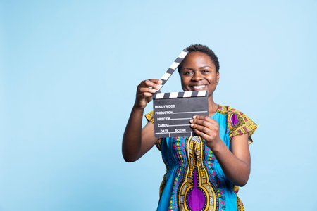African american girl posing with clapperboard in front of camera, smiling and using film slate in the post production movie industry. Female model working in filmography or videography.の写真素材