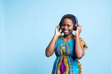 African american person having fun listening to songs on headphones, dancing around in the studio. Female model in traditional clothes enjoying stereo music on audio headset, modern mp3 tunes.の写真素材