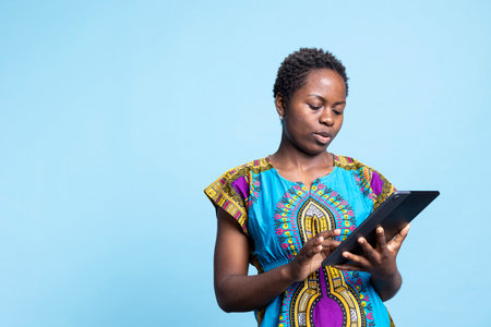 African american modern girl feels satisfied using a tablet over blue background, browsing online websites on mobile gadget. Young adult holds electronic device to navigate the internet.の写真素材