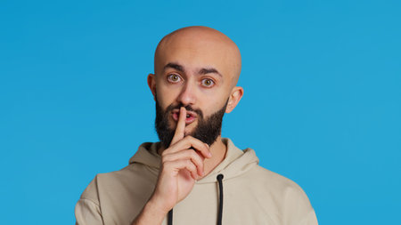 Middle eastern man doing hush silence symbol in studio, asking to keep a secret over blue background. Arab person creating mute gesture to be quiet in front of camera. Camera 2.の写真素材