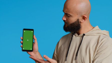 Muslim guy holding smartphone with greenscreen display, pointing at isolated chromakey template on mobile phone app. Young adult presenting blank copyspace mockup layout. Camera 2.の写真素材