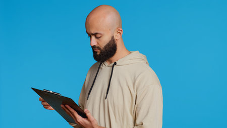 Middle eastern adult creating a checklist on clipboard papers, being focused on trying to plan tasks on an organizer in studio. Modern person looking at files and listing things to do. Camera 2.の写真素材