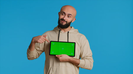 Arab guy holding device with greenscreen blank display, presenting isolated chromakey layout on tablet. Middle eastern adult using gadget to create promotional ad with copyspace. Camera 1.の写真素材