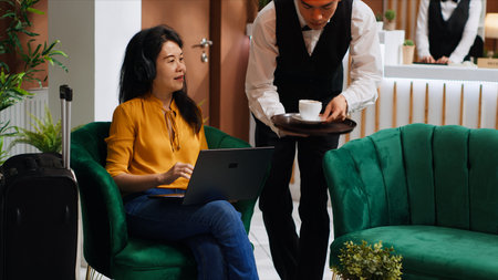 Asian woman receiving coffee while she works on laptop, sitting in lounge area at five star hotel and waiting for registration. Waiter serving drinks for guest relaxing, hospitality industry.の写真素材