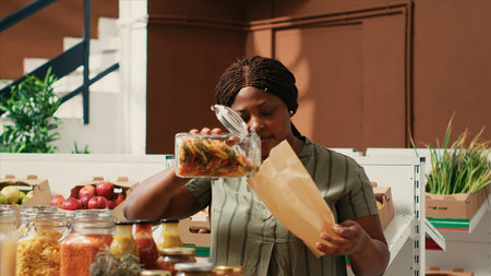 Woman client filling in paper bag with pasta sold as bulk item, buying various types of organic additives free products from local farmers market. Buyer searching for bio pantry supplies.の写真素材