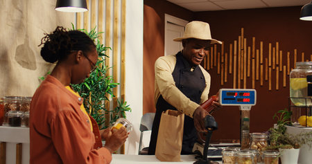 African american woman bringing natural products at checkout, talking to vendor about healthy eating while he scans goods. Local farmer selling his homegrown veggies and homemade items. Camera 2.の写真素材