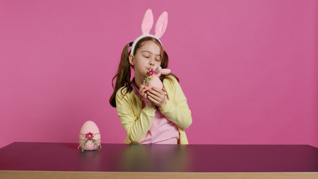 Smiling toddler with bunny ears showing her handmade easter ornaments, presenting a handcrafted decorated egg and rabbit. Young sweet schoolgirl holding festive decorations. Camera B.の写真素材