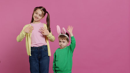 Joyful little kids waving in front of camera during easter holiday, smiling and wearing bunny ears. Brother and sister toddlers greeting someone in studio, adorable happy children. Camera A.の写真素材