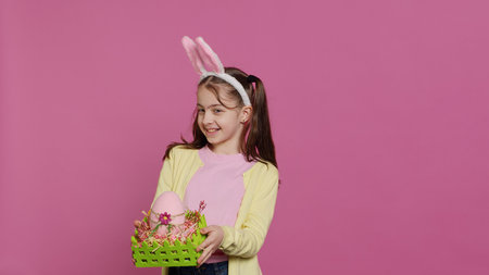 Smiling pleased girl presenting her handcrafted easter basket filled with painted eggs and other festive decorations for holiday celebration. Joyful small kid proud of her arrangement. Camera A.の写真素材