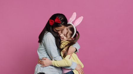 Lovely schoolgirl with bunny ears an her mom waving on camera, having fun and laughing against pink background. Cheerful mother and her daughter embracing and kissing each other. Camera A.の写真素材
