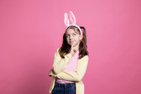 Small cheerful girl acting thoughtful about easter gifts in studio, thinking about holiday celebration with her bunny ears. Sweet young child with pigtails feeling confident in front of camera.の写真素材