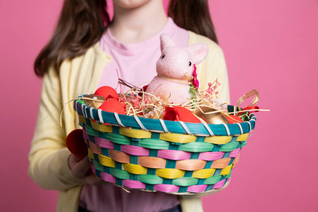 Small child presenting easter decorations and toys in a basket, holding colorful spring festive arrangements in front of camera. Young positive girl smiling in studio with adorable decor. Close up.の写真素材