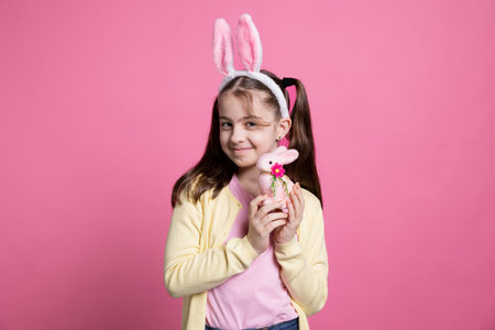 Positive cheerful girl with bunny ears posing over pink background, feeling enthusiastic about easter festive celebration. Cute small child holding a pink rabbit toy in front of camera, fluffy items.の写真素材
