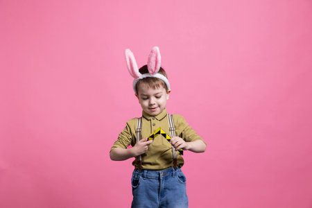 Joyful small child playing with a colorful toy in front of camera, enjoying leisure activity against pink background. Young cheerful boy wearing bunny ears and posing for an easter photoshoot.の写真素材