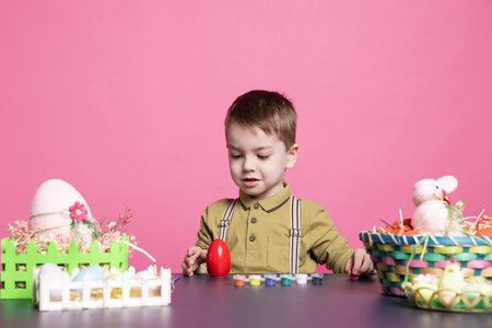 Lovely small boy decorating eggs with watercolors and imprints while making gorgeous ornaments for Easter Sunday event. Adorable youngster enjoys coloring with artistic supplies in the studio.の写真素材
