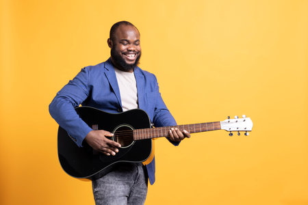 Smiling singer holding guitar, performing blues tunes during concert isolated over yellow studio background. Merry musician performing composition on stringed musical instrumentの写真素材
