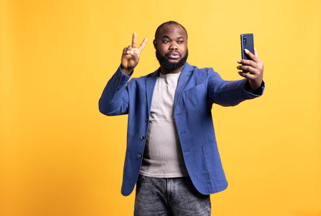 Man using smartphone to take selfies, doing victory hand sign. African american person taking photos using phone selfie camera, showing peace symbol gesturing, studio backgroundの写真素材